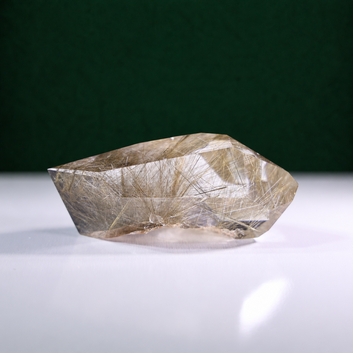 Polished rutilated quartz freeform, 185 g, main view on a white surface with a dark green background.