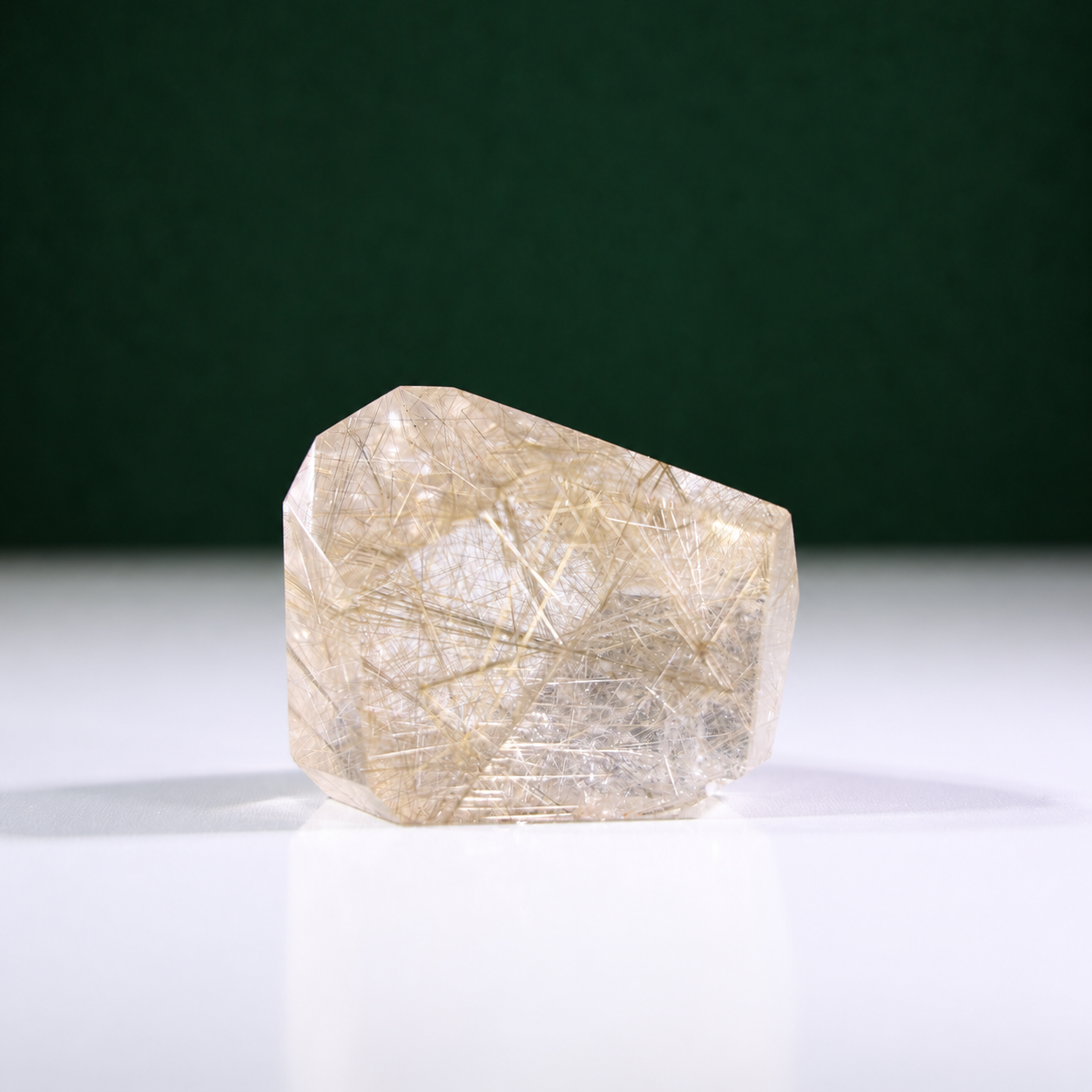Polished rutilated quartz freeform, 223 g, main view on a white surface with a dark green background.