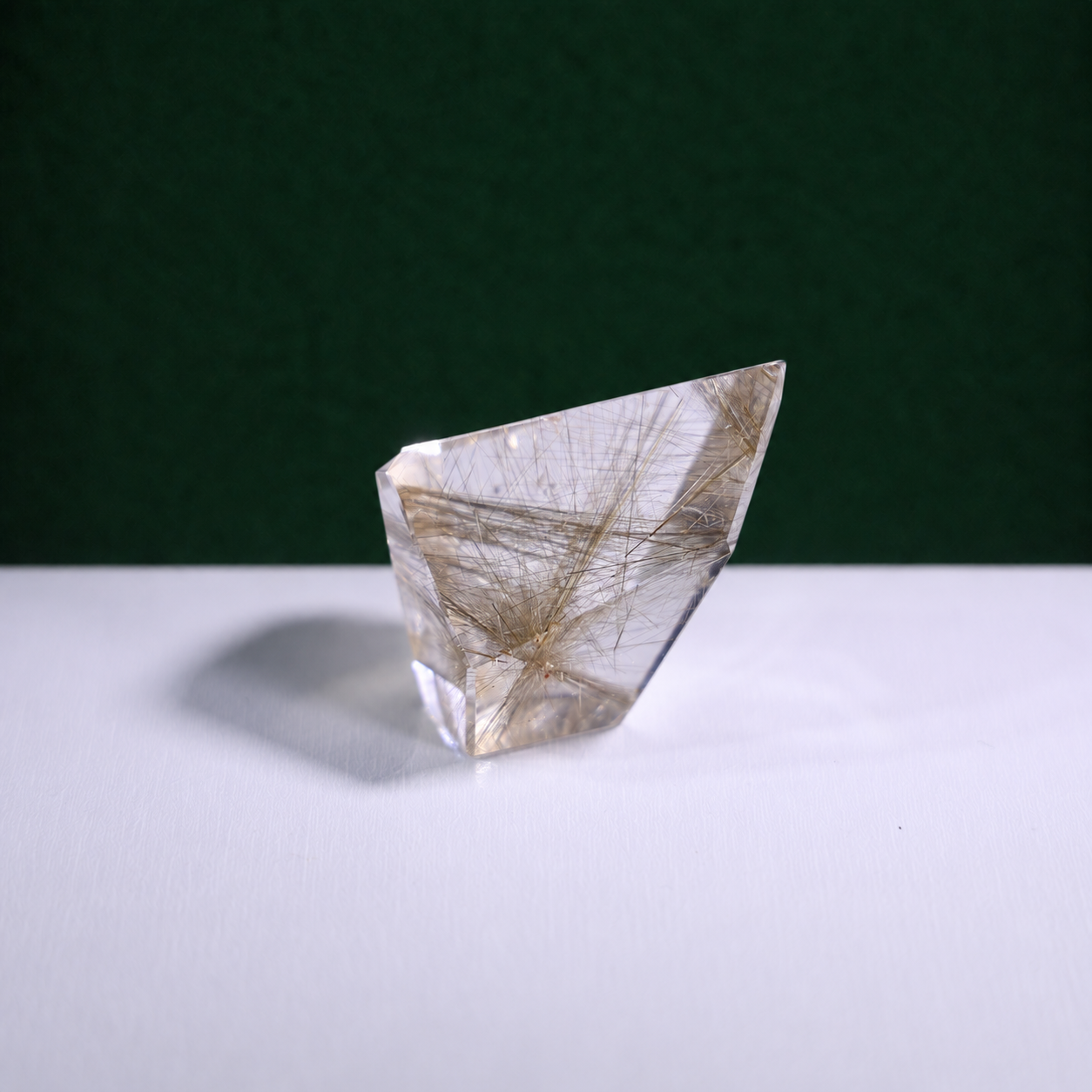 Polished rutilated quartz freeform, 62 g, main view on a white surface with a dark green background.