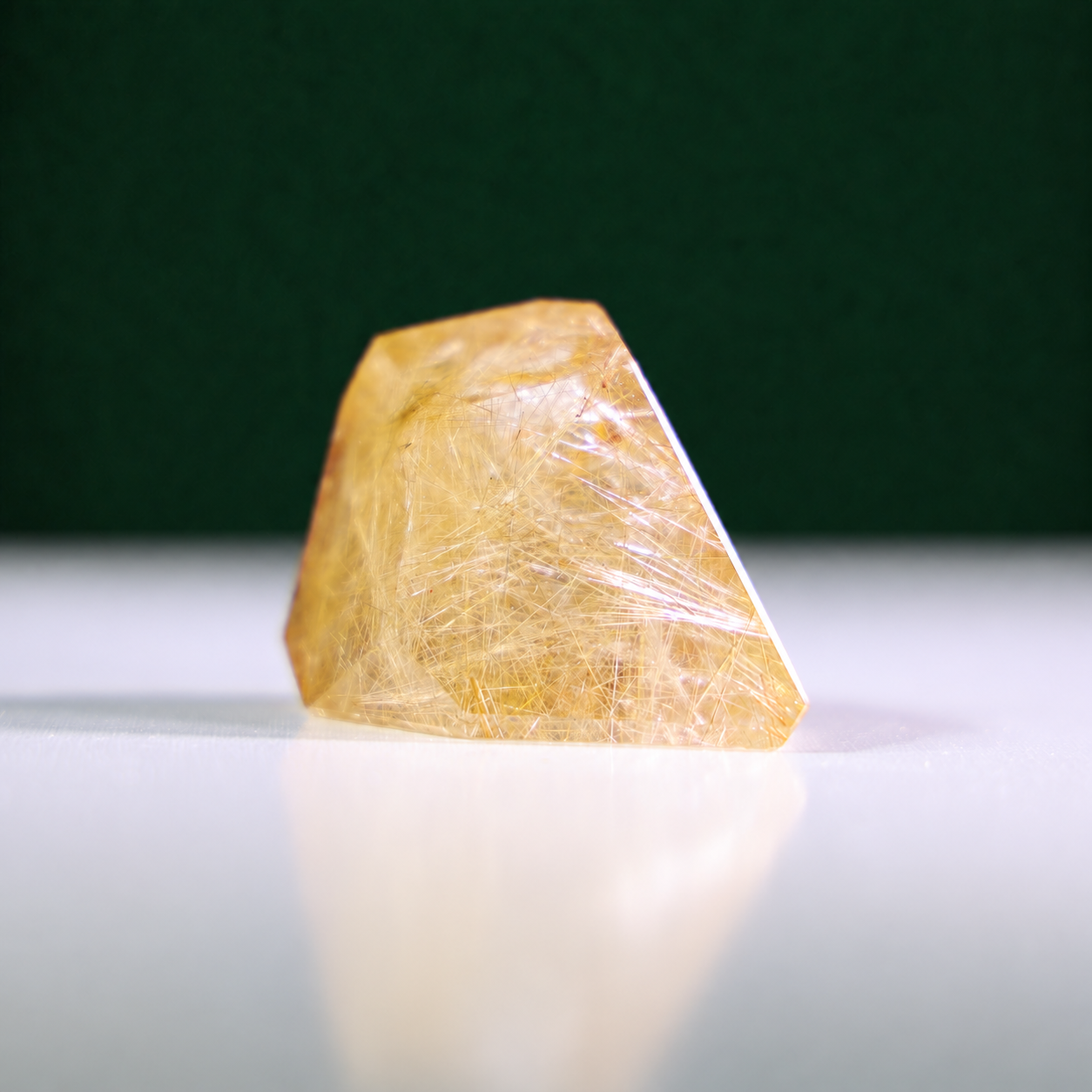 Polished rutilated quartz freeform, 75 g, main view on a white surface with a dark green background.